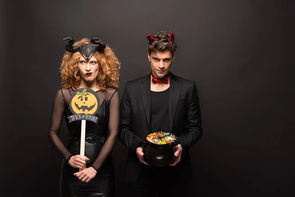 Couple réfléchi en costumes d'Halloween tenant pot avec des bonbons et citrouille truc ou traiter signe sur noir — Photo de stock