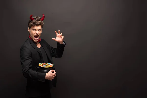 Sexy effrayant homme dans Halloween diable costume tenant pot avec des bonbons sur noir — Photo de stock