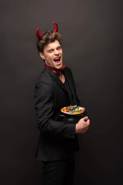 Bel homme en costume diable halloween tenant pot avec des bonbons sur noir — Photo de stock