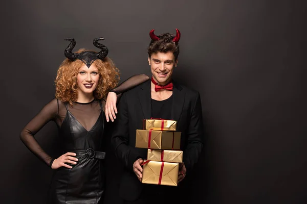 Hermosa pareja sonriente posando en trajes de halloween con regalos en negro - foto de stock