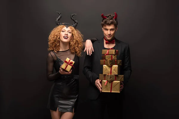 Beau couple sexy posant en costumes d'Halloween avec des boîtes-cadeaux sur noir — Photo de stock