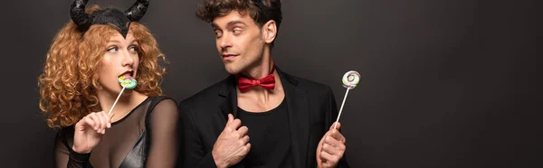 Beautiful young couple in halloween costumes holding lollipops on black — Stock Photo