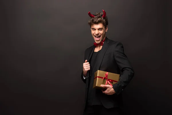 Guapo excitado hombre en traje de diablo celebración de cajas de regalo para halloween en negro - foto de stock