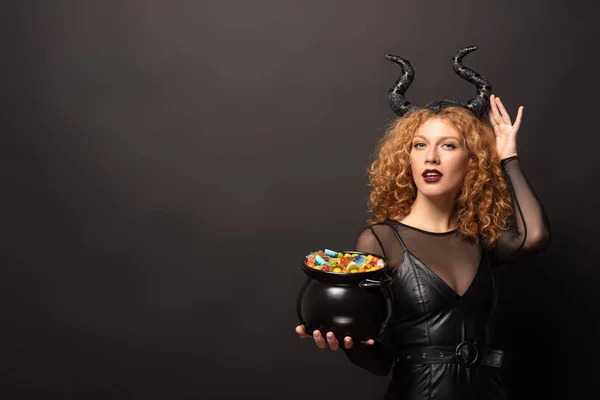 Lockige rothaarige Frau im bösartigen Kostüm, die einen Topf mit Bonbons für Halloween auf schwarz hält — Stockfoto