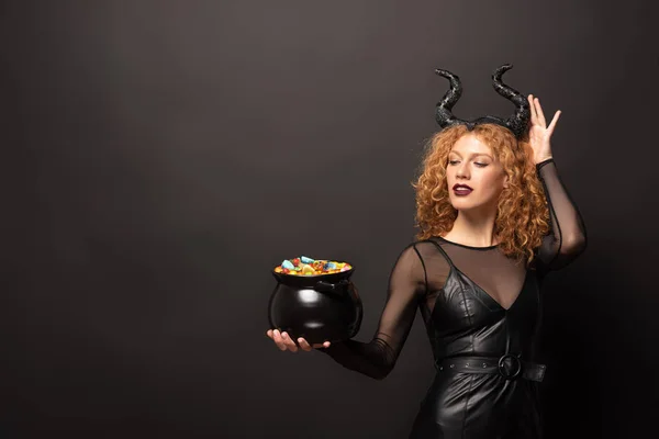 Atractiva mujer en traje maléfico sosteniendo olla con caramelos para halloween en negro - foto de stock