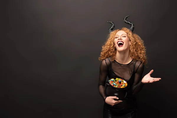 Laughing woman in maleficent costume holding pot with candies for halloween on black — Stock Photo