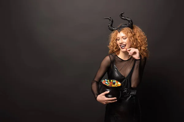 Красива кучерява жінка в чоловічому костюмі тримає горщик з цукерками для Хеллоуїна на чорному — стокове фото