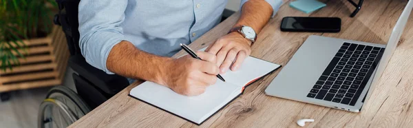 Vista recortada del hombre en silla de ruedas escribiendo en cuaderno en el lugar de trabajo, plano panorámico - foto de stock