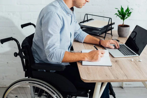 Vista recortada del hombre en silla de ruedas escribiendo en cuaderno y utilizando el ordenador portátil en el lugar de trabajo - foto de stock
