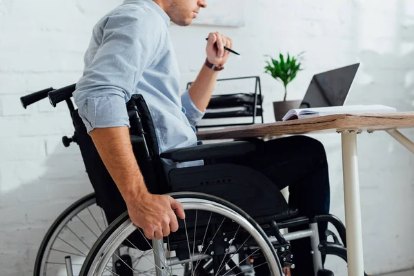 Обрезанный вид человека, сидящего в инвалидной коляске за ноутбуком на рабочем месте — стоковое фото