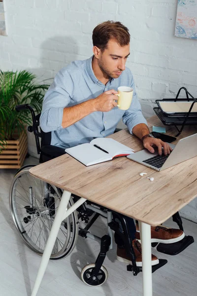 Man in wheelchair using laptop and drinking coffee at workplace — Stock Photo