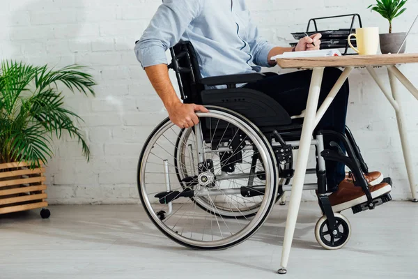 Vista recortada del hombre en silla de ruedas escribiendo en cuaderno en el lugar de trabajo - foto de stock