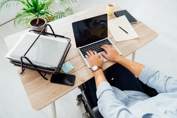 Vista recortada del hombre en silla de ruedas usando portátil con pantalla en blanco en el lugar de trabajo — Stock Photo