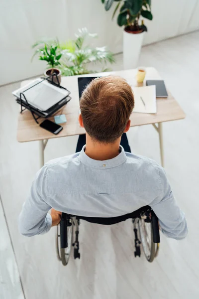 Rear view of man in wheelchair sitting at desk with laptop and papers — Stock Photo