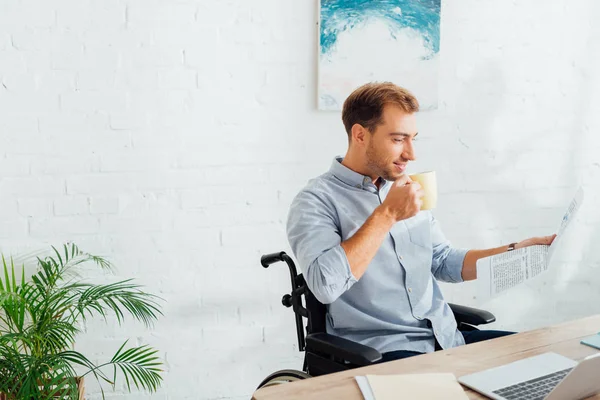 Uomo sorridente sulla sedia a rotelle che beve tè e legge giornali alla scrivania — Foto stock