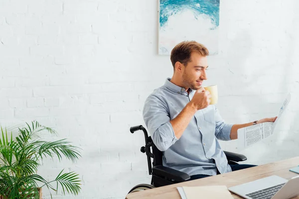 Lächelnder Mann im Rollstuhl trinkt Tee und liest Zeitung am Schreibtisch — Stockfoto