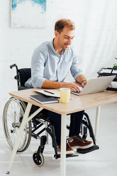 Hombre de la felicidad en silla de ruedas escribiendo en el teclado portátil en el lugar de trabajo - foto de stock