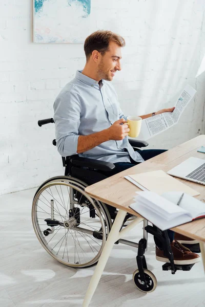Felicidad hombre en silla de ruedas beber té y leer el periódico en el escritorio - foto de stock