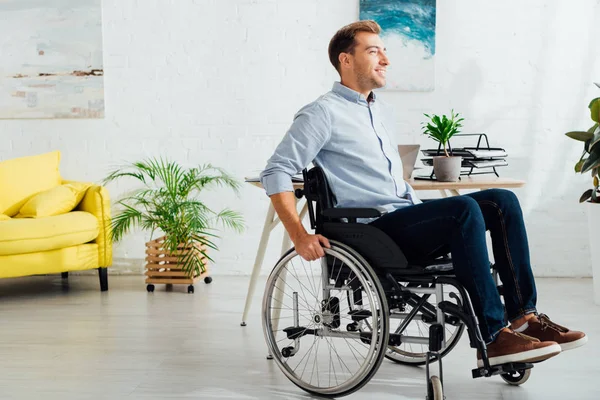 Hombre sonriente sentado en silla de ruedas y mirando hacia otro lado en la sala de estar - foto de stock