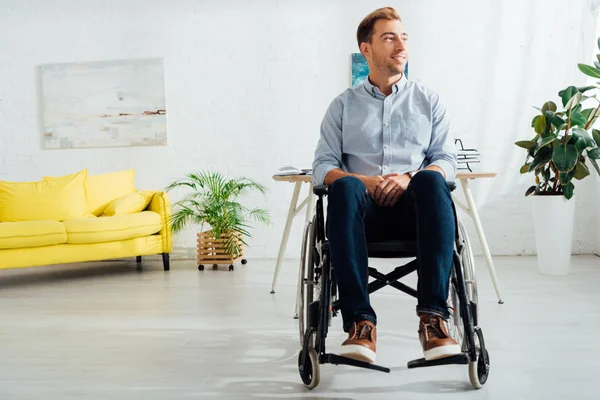 Улыбающийся мужчина сидит в инвалидной коляске и смотрит в сторону в гостиной — стоковое фото