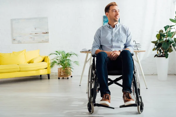 Hombre sonriente sentado en silla de ruedas y mirando hacia otro lado en la sala de estar - foto de stock