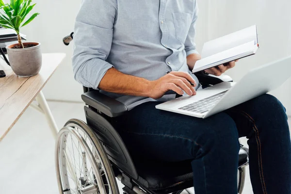 Cropped view of man in wheelchair holding notebook and laptop — Stock Photo