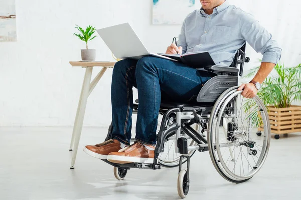 Vue recadrée de l'homme en fauteuil roulant écrit dans un cahier et tenant un ordinateur portable — Photo de stock