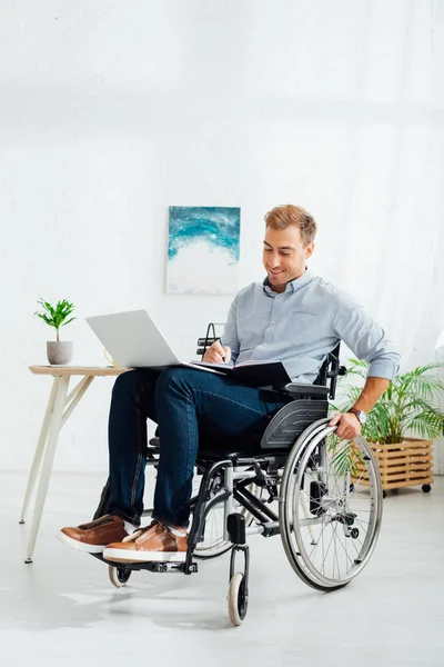 Hombre sonriente en silla de ruedas escribiendo en un cuaderno y sosteniendo el portátil en la sala de estar - foto de stock