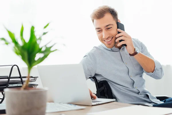 Smiling freelancer talking on smartphone and using laptop at desk — Stock Photo