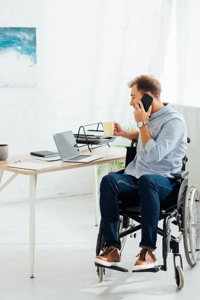 Uomo in sedia a rotelle che parla su smartphone e tiene in mano una tazza di caffè sul posto di lavoro — Foto stock