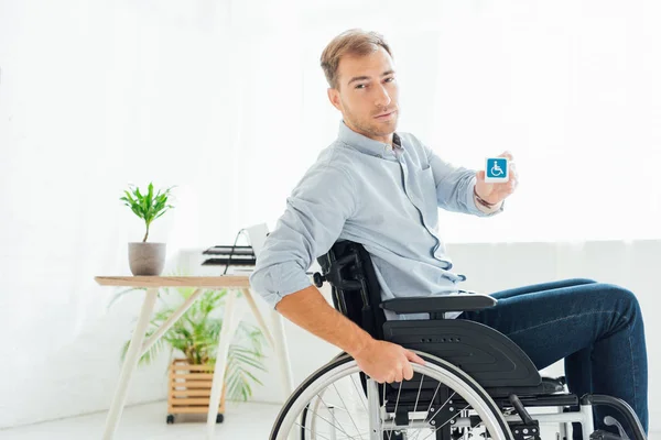 Человек в инвалидном кресле показывает куб с вывеской для инвалидов и смотрит в камеру — стоковое фото