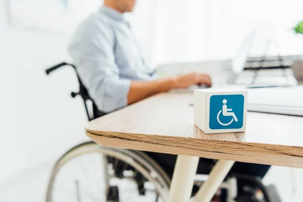 Enfoque selectivo del cubo con signo de discapacidad y el hombre en silla de ruedas sentado en la mesa - foto de stock