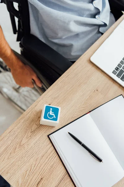 Cube with disabled sign and man in wheelchair sitting at table with laptop and notebook — Stock Photo