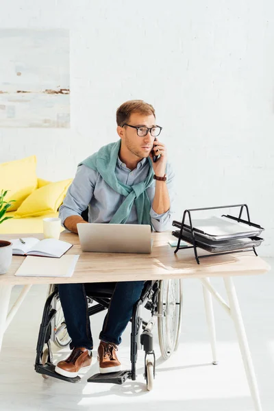 Hombre en silla de ruedas hablando en el teléfono inteligente mientras trabaja en el portátil - foto de stock