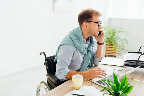 Casual businessman in wheelchair talking on smartphone and using laptop at desk — Stock Photo