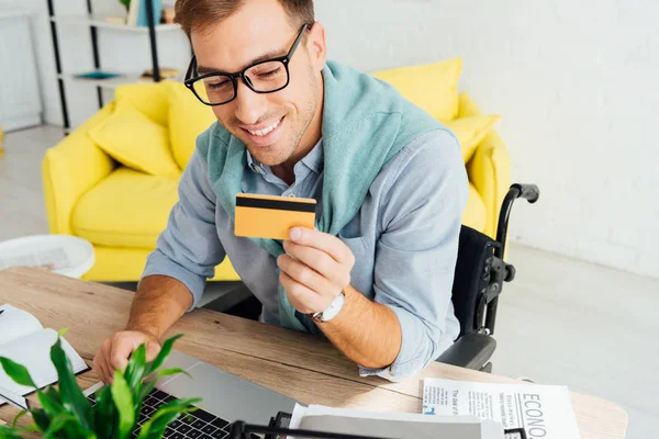 Улыбающийся человек в инвалидной коляске смотрит на кредитную карту и использует ноутбук за столом — стоковое фото