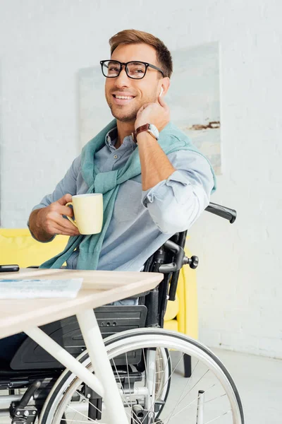 Uomo sorridente in sedia a rotelle utilizzando auricolari wireless e tenendo tazza di tè — Foto stock