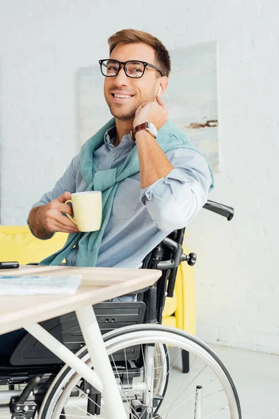 Hombre sonriente en silla de ruedas usando auriculares inalámbricos y sosteniendo la taza de té - foto de stock
