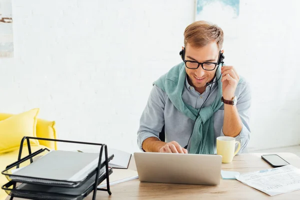 Hombre sonriente en gafas con auriculares y portátil en la mesa de trabajo - foto de stock