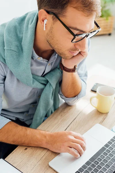 Freelancer con la mano en la barbilla utilizando auriculares inalámbricos y portátil en el escritorio de trabajo - foto de stock