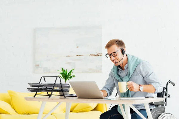 Uomo in sedia a rotelle con auricolare, lavorando sul computer portatile e tenendo tazza in soggiorno — Foto stock