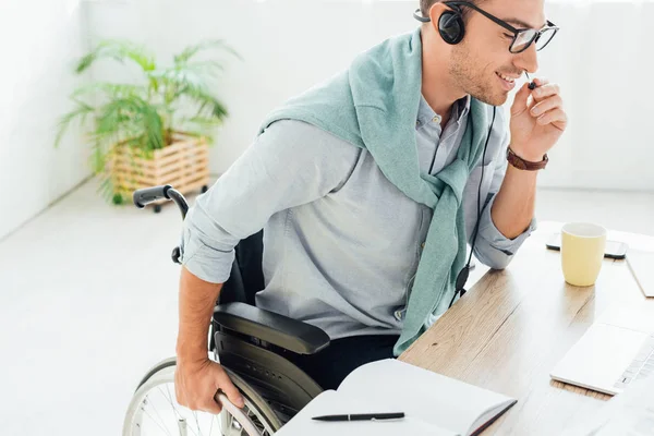 Operador de centro de llamadas sonriente en silla de ruedas hablando con auriculares - foto de stock