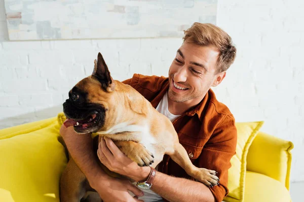 Sorrindo homem abraçando bulldog francês no sofá — Fotografia de Stock