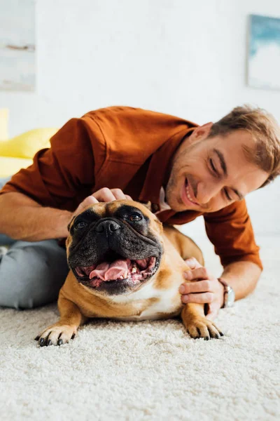 Uomo sorridente che gioca con bulldog francese su tappeto in soggiorno — Foto stock