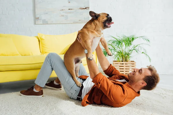 Vista lateral del hombre jugando con bulldog francés en la alfombra en casa - foto de stock