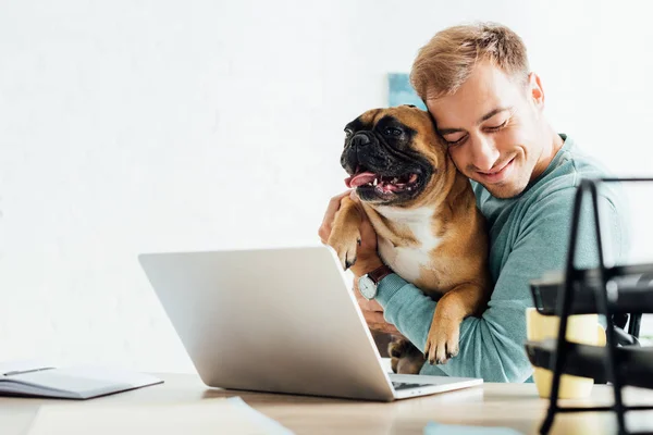 Sorrindo homem abraçando bulldog francês enquanto trabalhava no laptop — Fotografia de Stock
