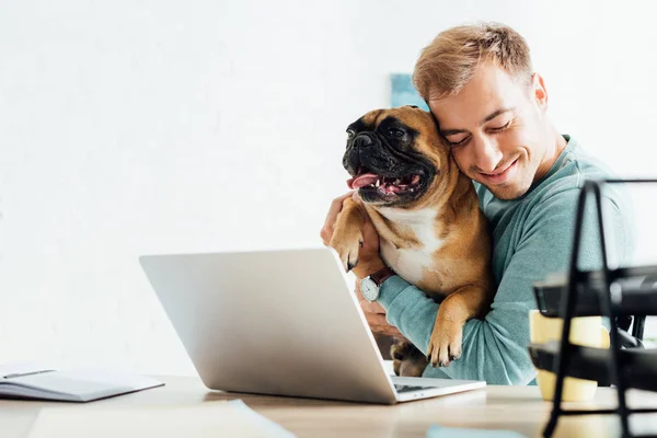 Homme souriant étreignant bouledogue français tout en travaillant sur ordinateur portable — Photo de stock