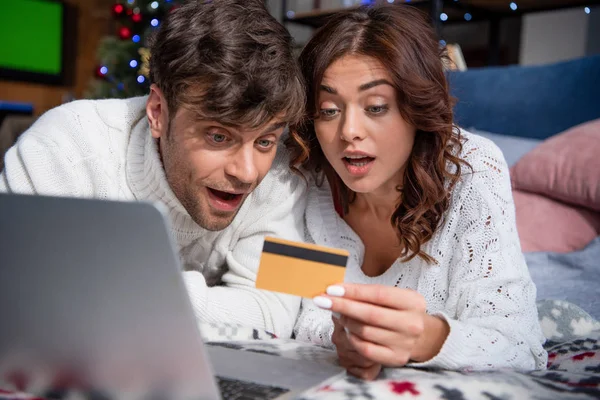 Shocked girlfriend and handsome boyfriend in sweaters looking at credit card — Stock Photo
