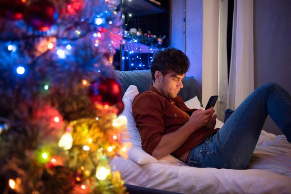 Foco seletivo de namorado bonito sentado na cama e usando smartphone no Natal — Fotografia de Stock