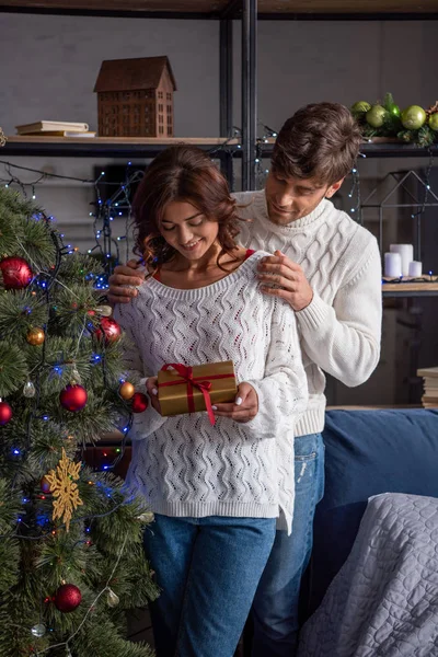 Улыбающийся парень обнимает девушку в свитере с подарком на Рождество — стоковое фото